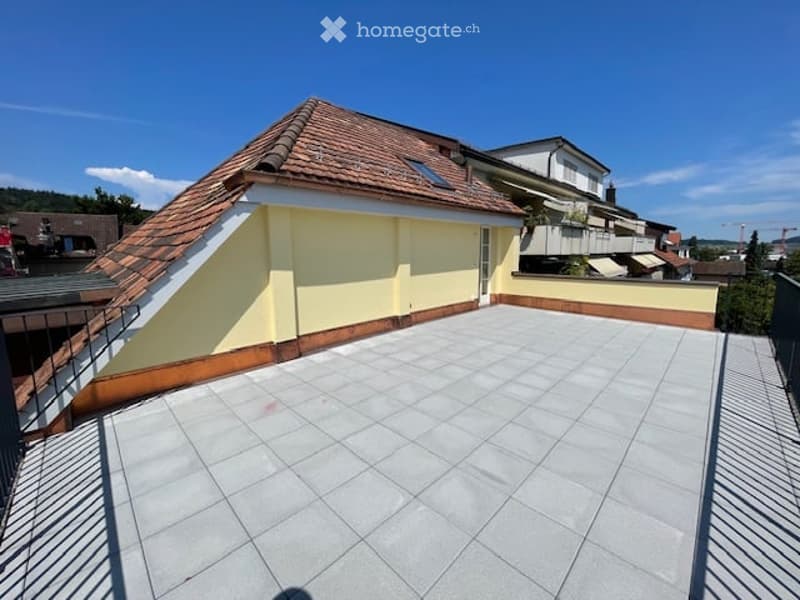 Roof terrace with tiled flooring, modern railing, and a view of surrounding buildings and greenery.