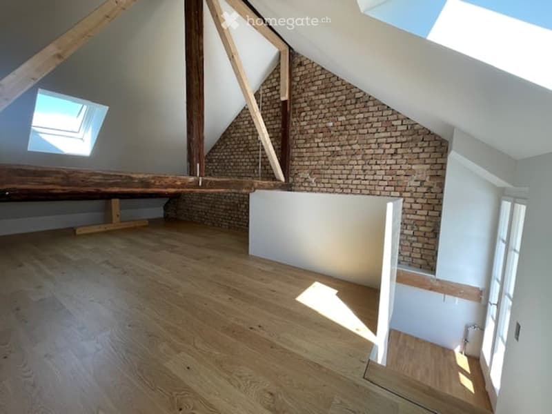Empty attic space with exposed wooden beams, brick wall, skylight window, and wooden flooring.