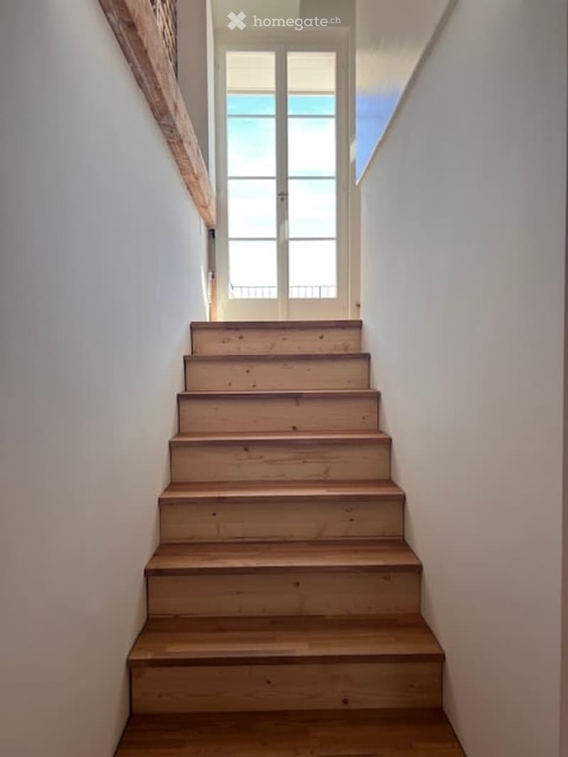 Wooden staircase leading up, white walls, a door at the top, and a window providing natural light.
