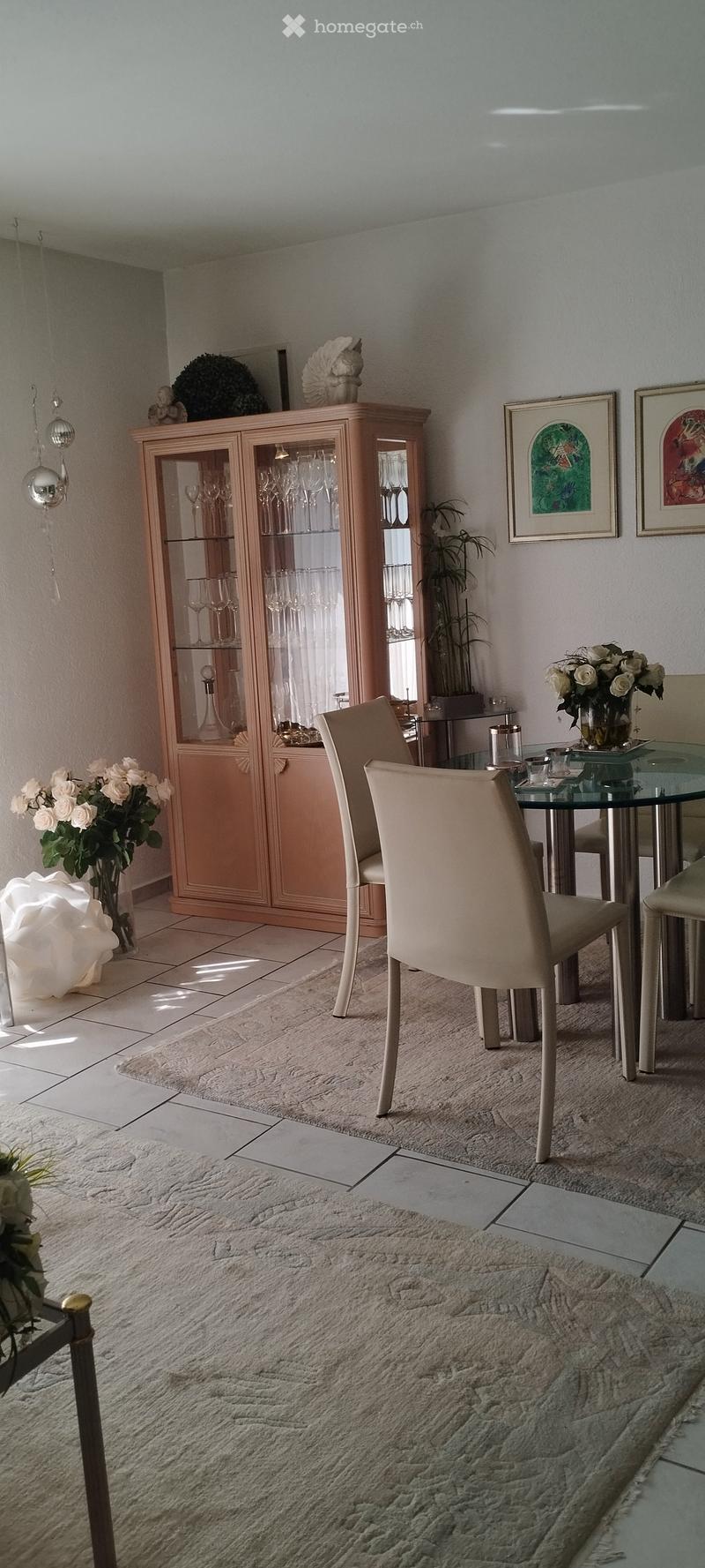 Dining room with a glass-top table, four beige chairs, a light wood cabinet with glassware, and decorative vases and artwork on the walls.