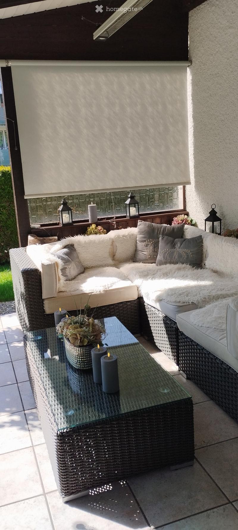A cozy outdoor seating area with a wicker sofa, plush cushions, a glass coffee table, candles, and decorative lanterns.