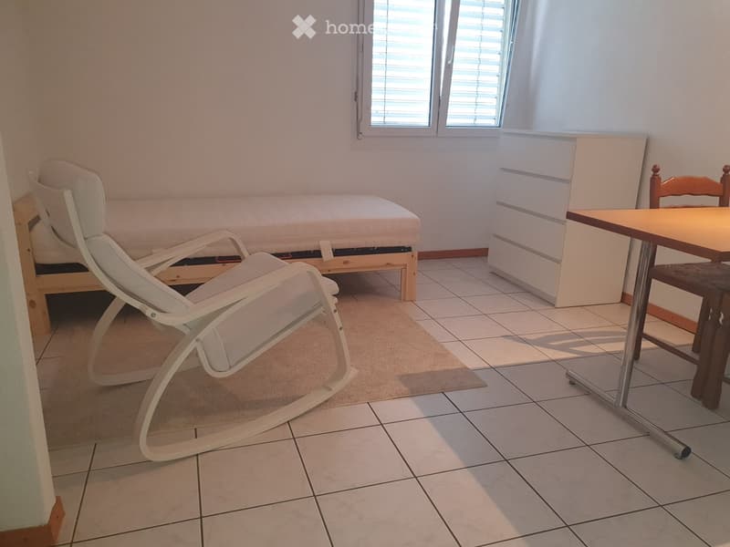A furnished room with a bed, a white lounge chair, a small table, and a rug on the floor.