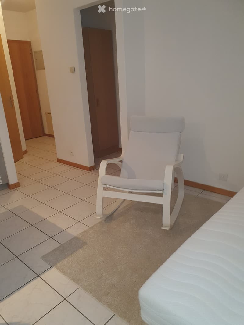 A small, well-lit room with tiled flooring, a light-colored armchair, a sofa, and a beige rug.