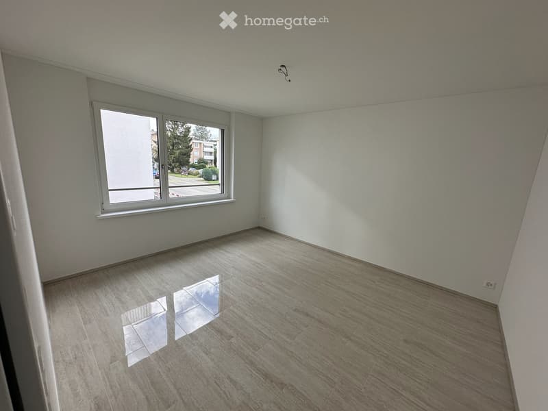 Empty room with light-colored walls and wooden flooring, featuring a large window with a view of the street.