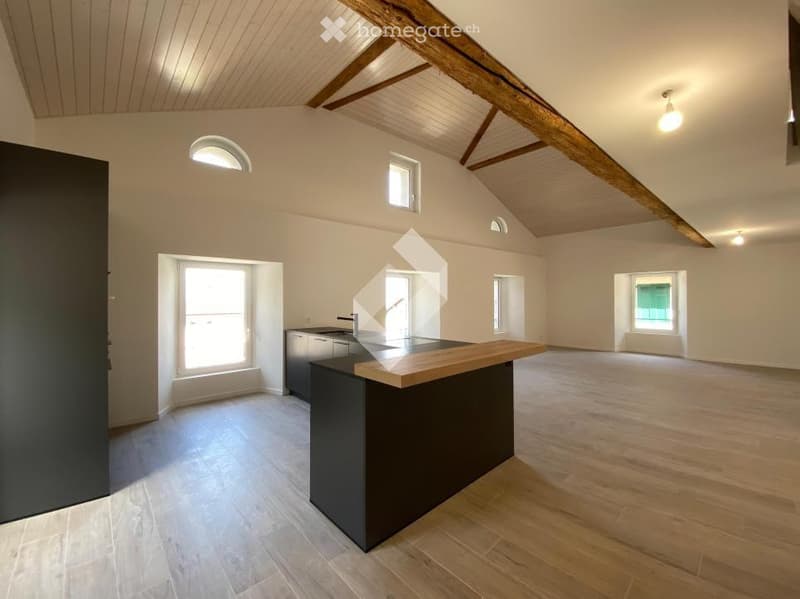 Open-plan kitchen and living area with exposed wooden beams, light wooden flooring, and large windows. Features a modern kitchen island with a sink and faucet.