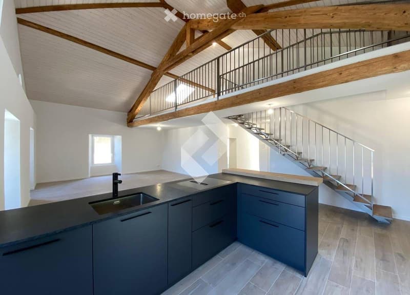 Modern kitchen with dark grey cabinets, a central island, and a staircase leading to an upper level. Exposed wooden beams on the ceiling.
