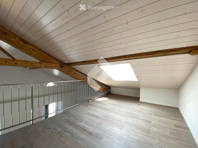 Empty attic space with wooden ceiling beams, exposed wooden beams, wooden floor, and a metal railing leading to a staircase. Natural light streams in from a skylight.