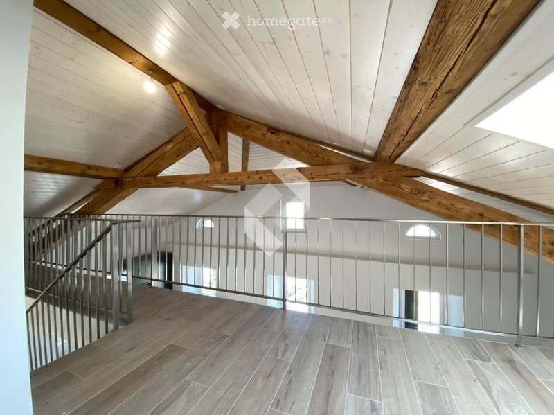 Spacious hallway with exposed wooden beams and a modern metal railing leading to an upper level. Light-colored wooden flooring and white walls.