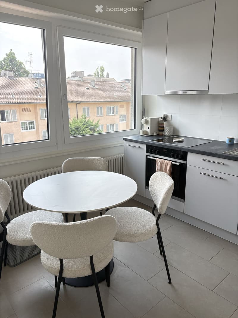 Modern kitchen with white cabinets, a round dining table with four chairs, and a built-in oven. Large windows offer a view of the city.