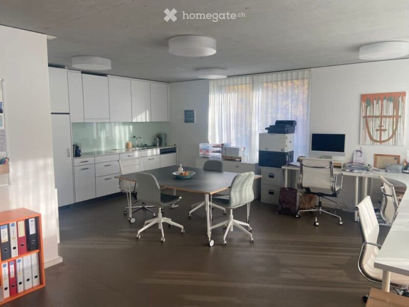 The image shows a modern, well-lit office space with a large wooden table surrounded by ergonomic chairs. There are white cabinets and a small sink area in the background, along with a computer setup on a desk. The room features large windows with sheer c