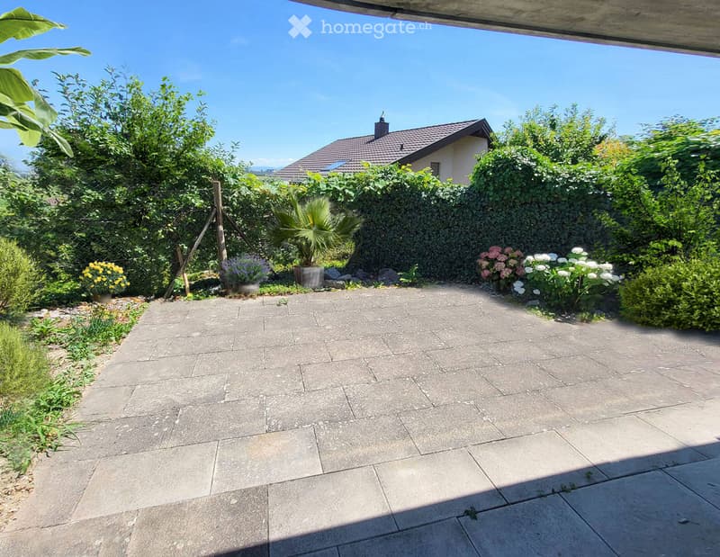 Garden area with a paved patio, surrounded by greenery, flowers, and a house in the background.