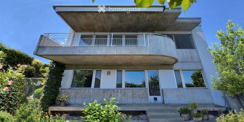 Modern two-story concrete house with large windows, a balcony, and a raised ground floor.
