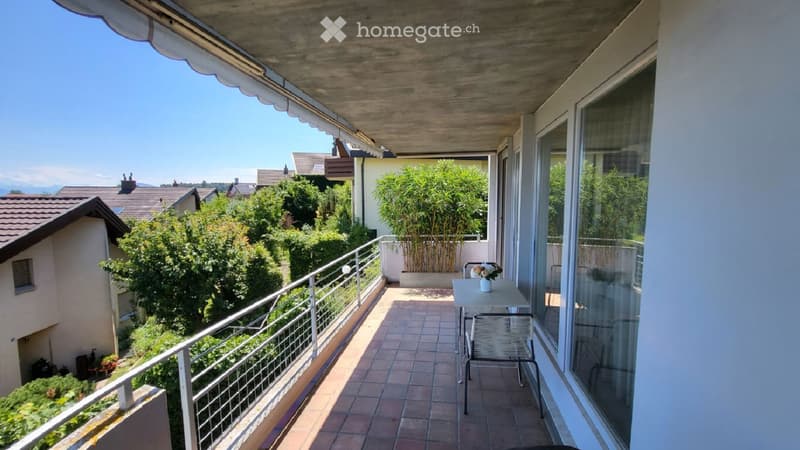 Balcony with tiled floor, metal railing, small table and chair, overlooking a residential area with greenery and houses.