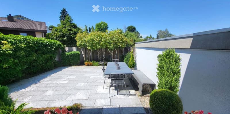 Modern patio with a stone-tiled floor, a rectangular table with four chairs, and surrounding greenery including shrubs and trees.