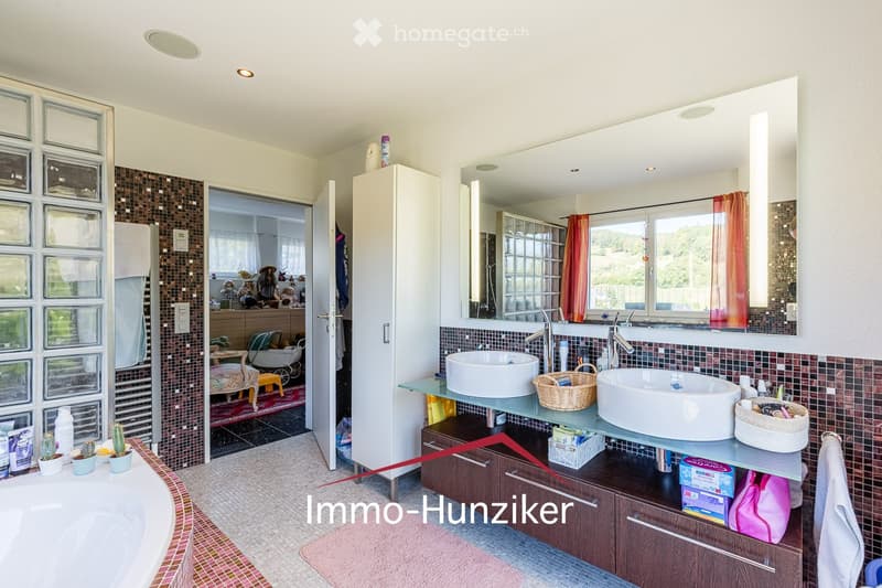 Modern bathroom with a bathtub, double sink, large mirror, and wooden cabinetry. Features mosaic tiles, a wicker basket on the counter, and a small seating area visible through an open doorway.