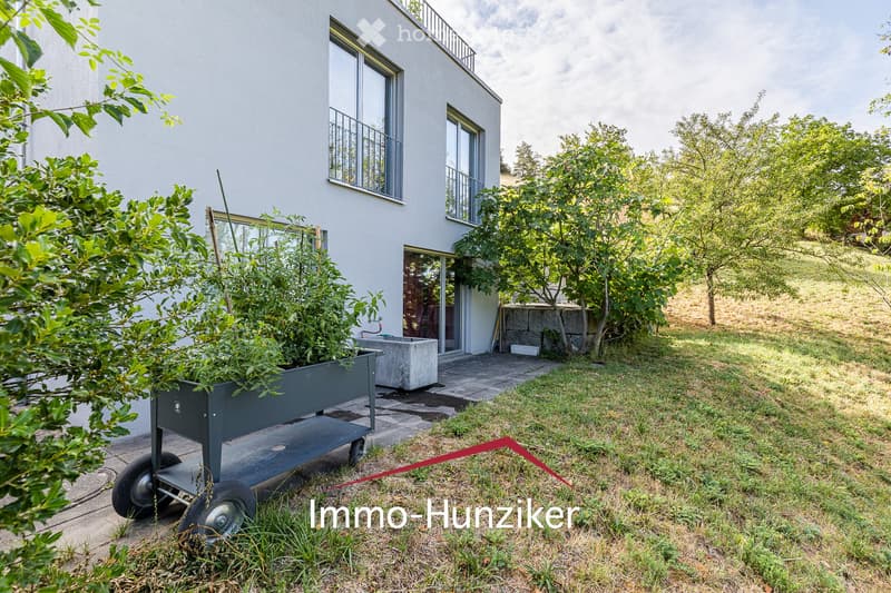 A modern house with a balcony, surrounded by a well-maintained garden with trees, shrubs, and a paved patio area.