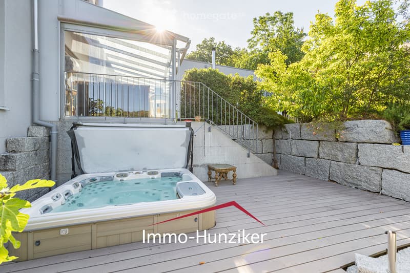 A wooden deck area with a hot tub, small table, and lush greenery, featuring a modern glass balcony with metal railing and stone wall.