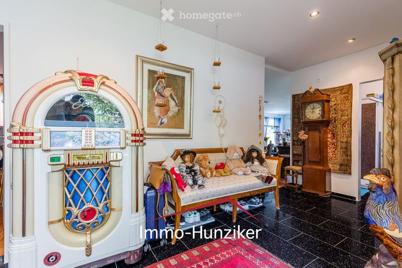 A cozy living room with a retro jukebox, a sofa adorned with stuffed animals, a framed painting, a wooden clock, and a small table with a stool nearby.