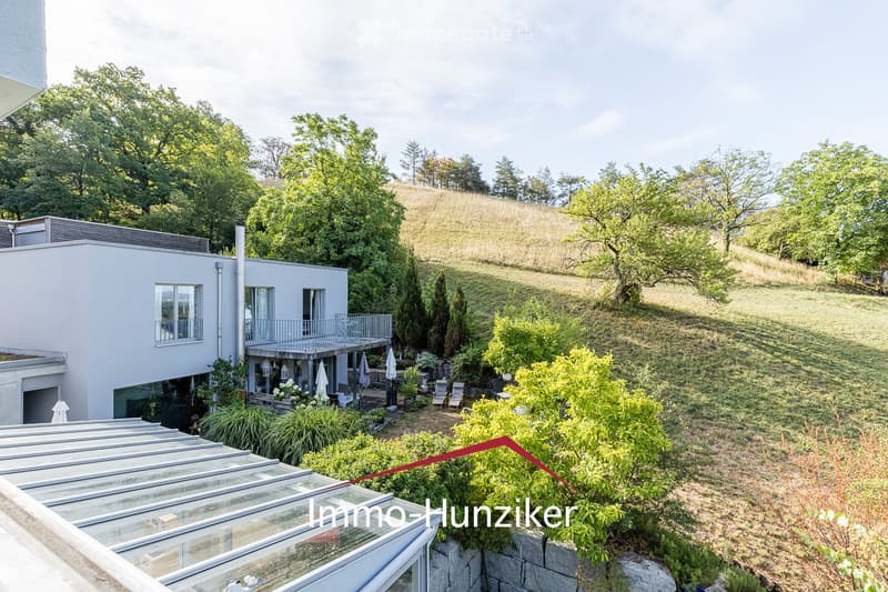 Modern two-story house with a flat roof, large windows, and a balcony. The surrounding area includes a well-maintained garden with various plants, trees, and outdoor seating.