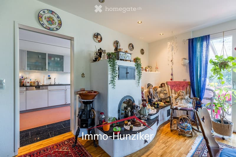 A cozy living room with a fireplace, wooden floor, and a kitchen area visible in the background. The room features a sofa, a small table with dolls and decorative items, a hanging plant, and a large window with blue curtains. The kitchen has white cabinet