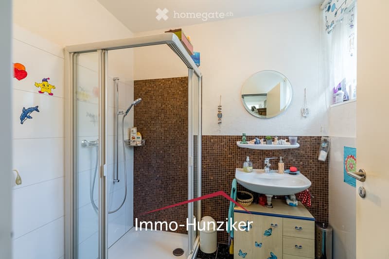Bathroom with a sink, mirror, shower, and various toiletries. Features a wooden cabinet with drawers and a small shelf above the sink.
