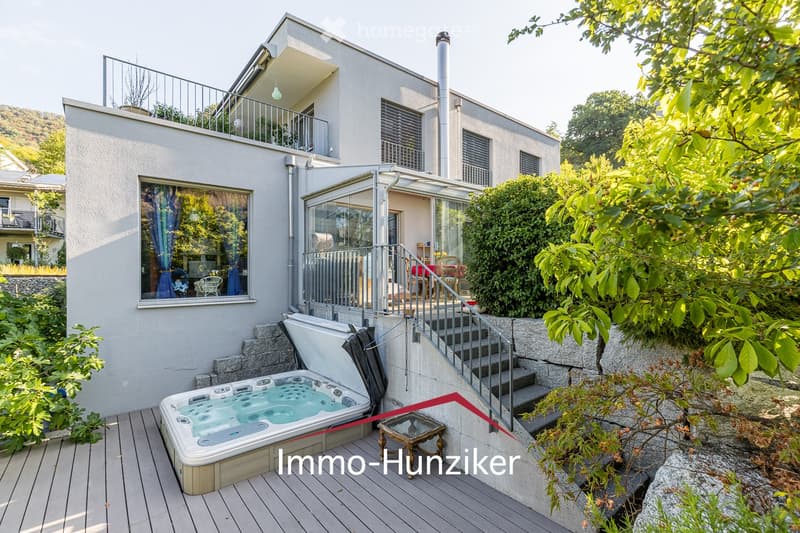 Modern house with a wooden deck, hot tub, small table, and stairs leading to a covered patio area with outdoor furniture.