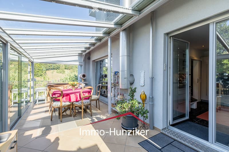 A spacious, covered balcony with large glass panels, tiled floor, and a dining setup including a table with a pink tablecloth and chairs. There are potted plants and a view of greenery outside.