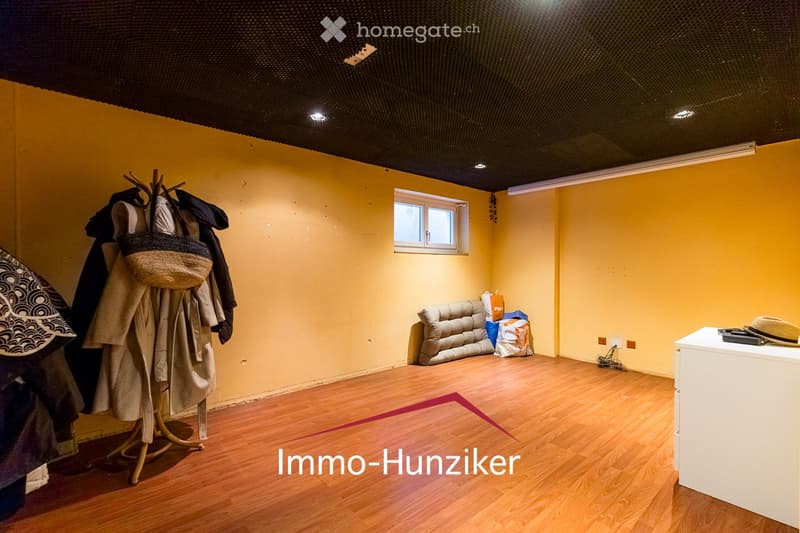 A small, furnished room with yellow walls, wooden flooring, a single window, a sofa, a coat rack with jackets and bags, and a small table with a hat on it.