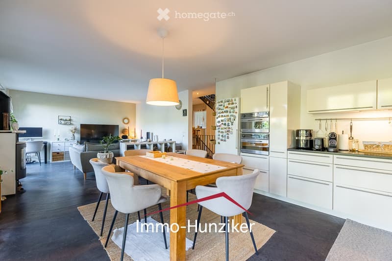 Open-plan living and dining area with a grey sofa, coffee table, dining table with chairs, and a modern kitchen featuring white cabinets, stainless steel appliances, and a coffee maker on the counter.
