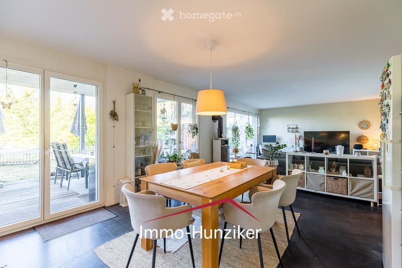Bright dining area with a wooden dining table, chairs, a cabinet, and a TV in the background. Large sliding glass doors lead to a balcony with outdoor furniture.