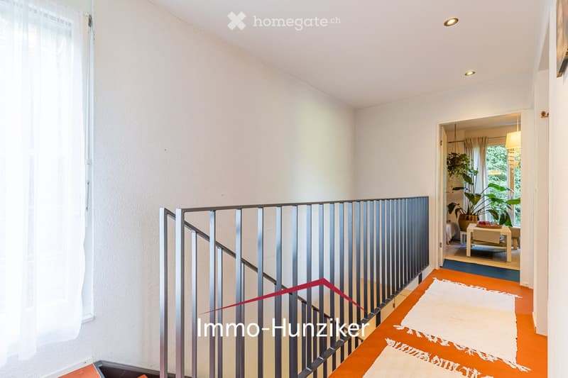 A modern hallway with a sleek metal railing leading to a room with visible furniture and plants.
