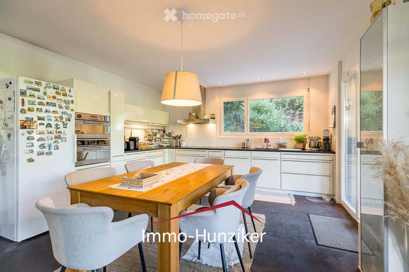 Modern kitchen with white cabinets, a central wooden dining table surrounded by chairs, a pendant light, and various appliances including a refrigerator, oven, coffee maker, and blender. Large windows provide natural light, and a small plant adds a touch