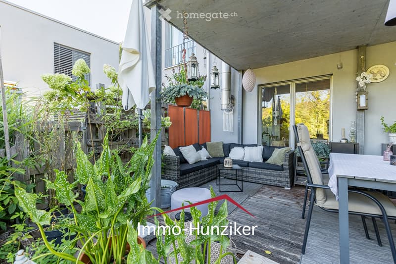 A modern, well-furnished balcony with a wooden deck, a black wicker sofa with cushions, a small coffee table, a white stool, a dining table with chairs, and several potted plants. The space is covered by a pergola with hanging lanterns and has a view of t