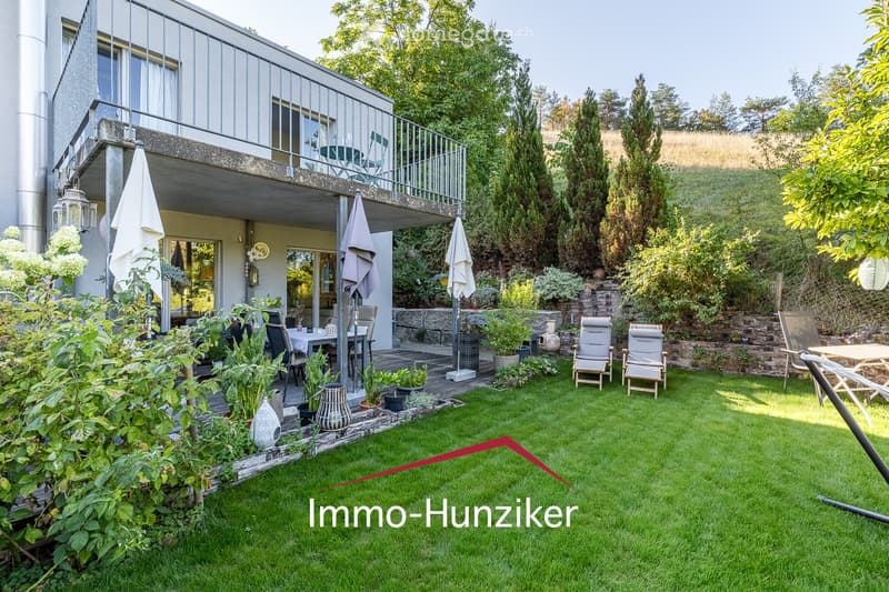 Well-maintained garden with a green lawn, patio seating area, outdoor furniture, and a modern house with a balcony and large windows.