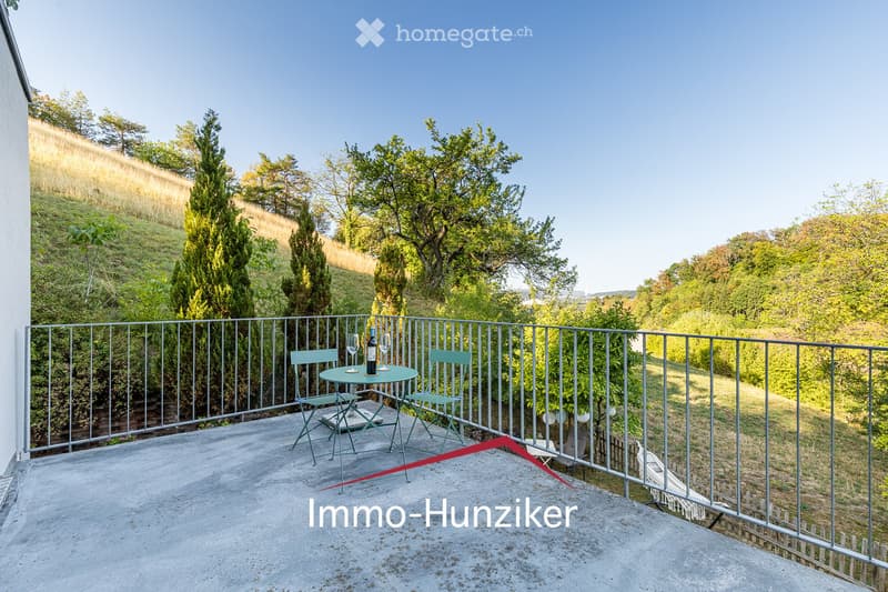 Balcony with a metal railing, a small round table with two chairs, a bottle, and two glasses, overlooking a lush green landscape.