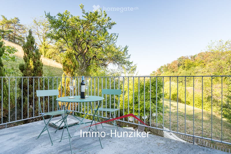 A small balcony or terrace with a round table, two chairs, a wine bottle, and two wine glasses, surrounded by a metal railing and lush greenery.