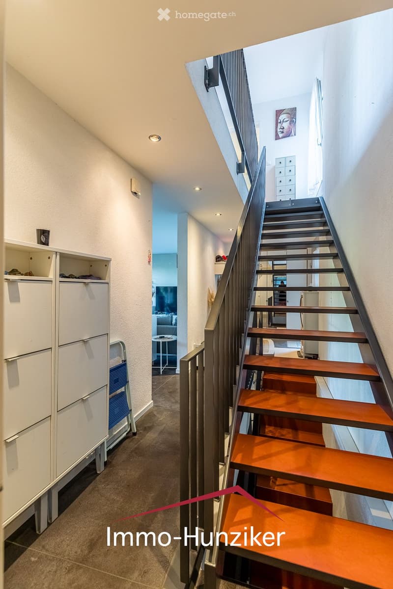 Modern hallway with a wooden staircase, white walls, and built-in storage units. The staircase features a metal railing and wooden steps, leading to another room visible in the background.