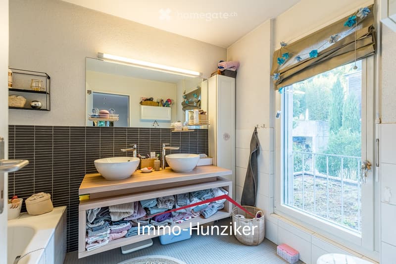 Modern bathroom with double sink vanity, large mirror, shelves, and towels neatly arranged. Features dark tiled walls and a window with blinds.