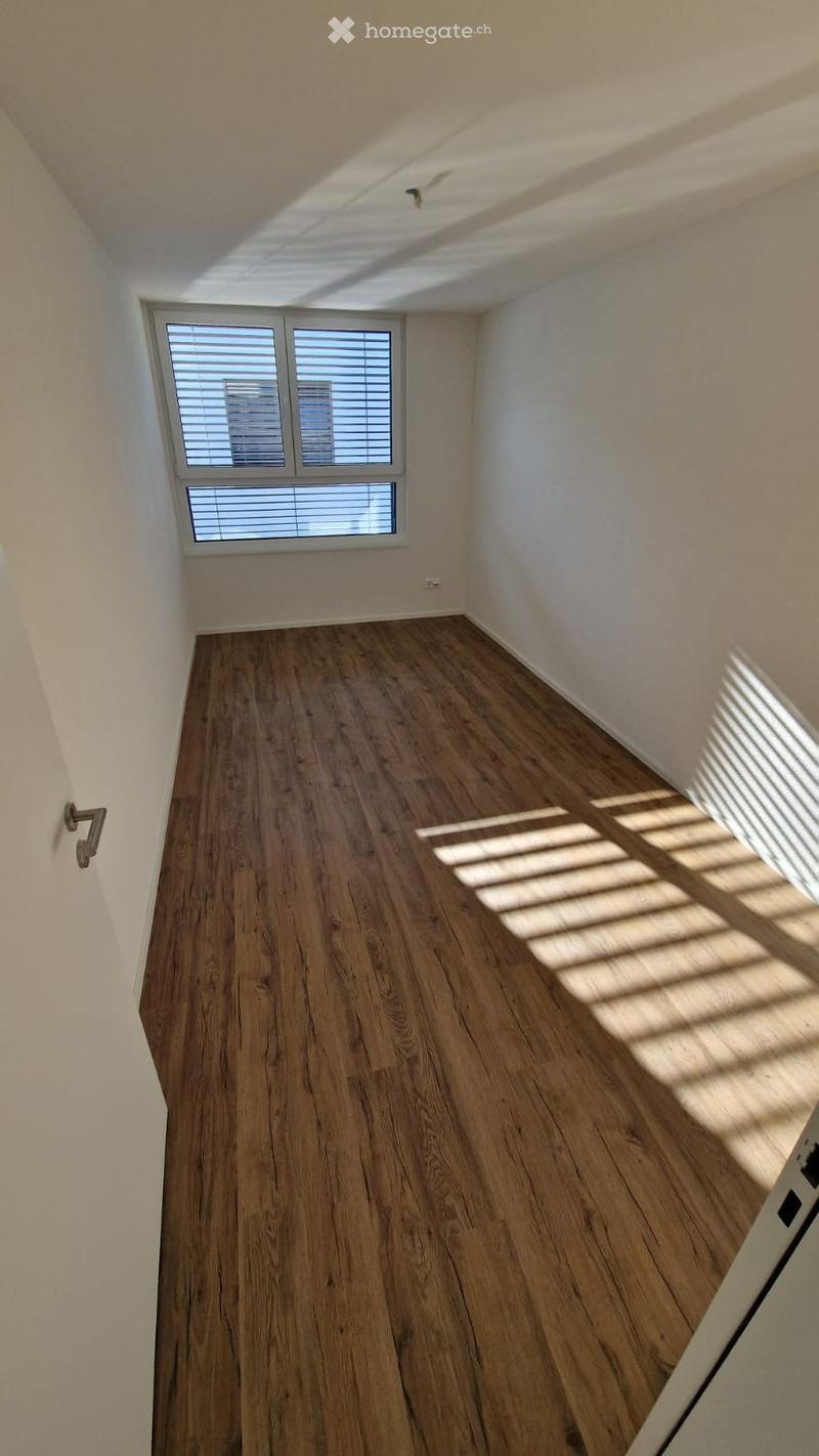 Empty room with wooden flooring and white walls, featuring a window with blinds and a door handle visible on the left side.