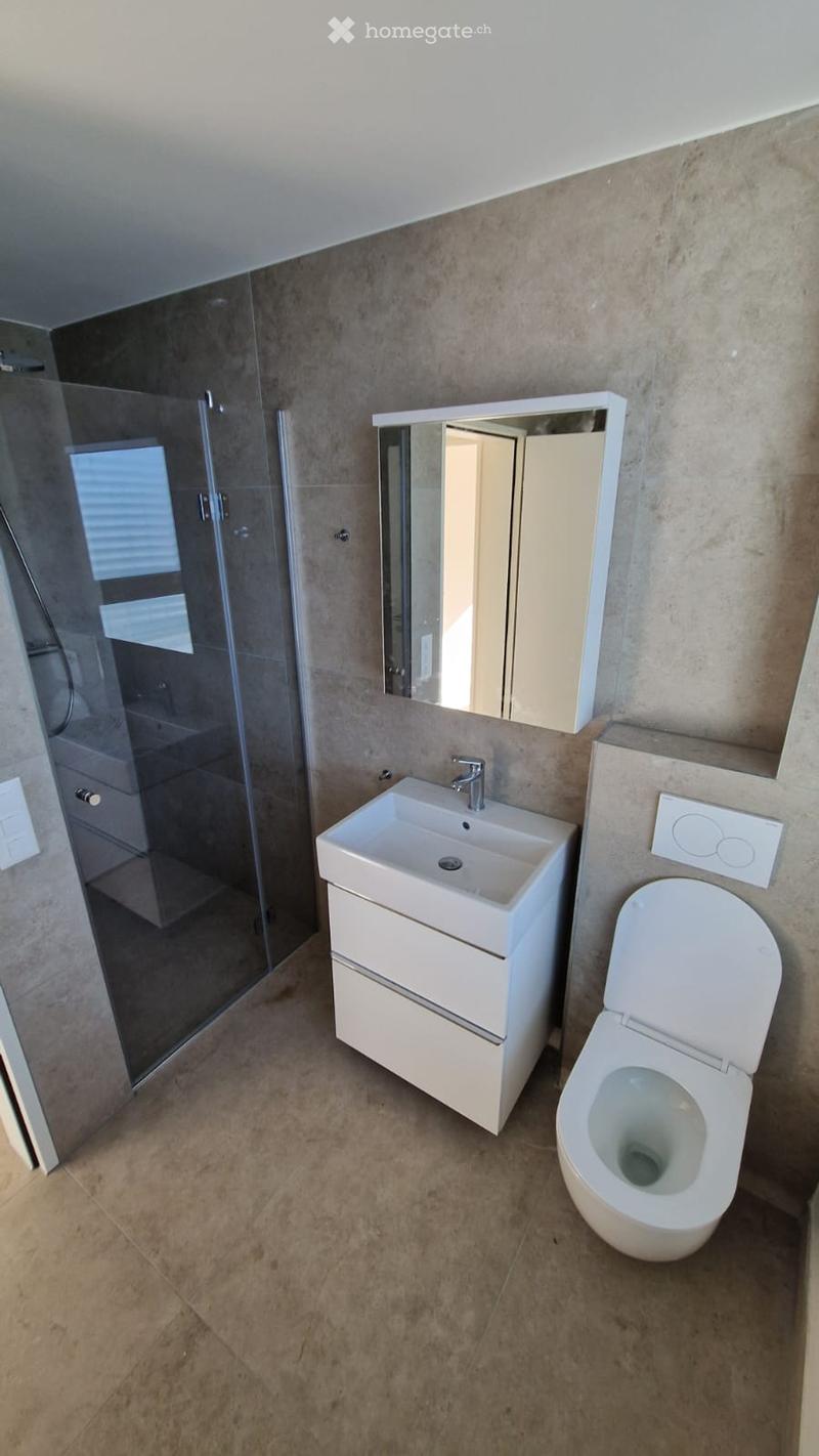Modern bathroom with a white sink, toilet, mirror, and glass-enclosed shower. The walls are tiled in a neutral tone, and the floor is made of beige tiles.