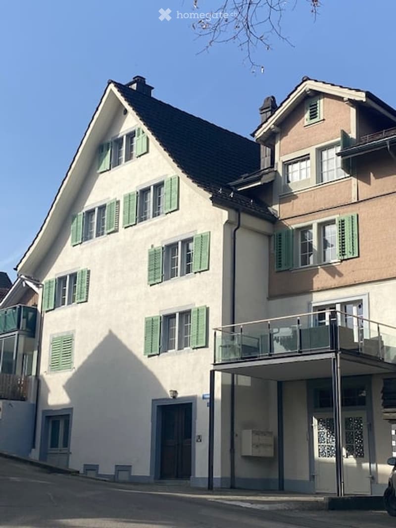 Two-story residential building with green shutters, white walls, and a balcony on the right side.
