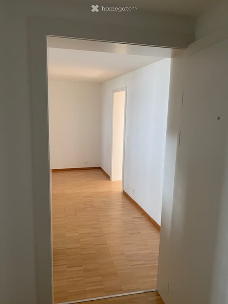 Empty room with light wooden flooring and white walls, visible through an open doorway.
