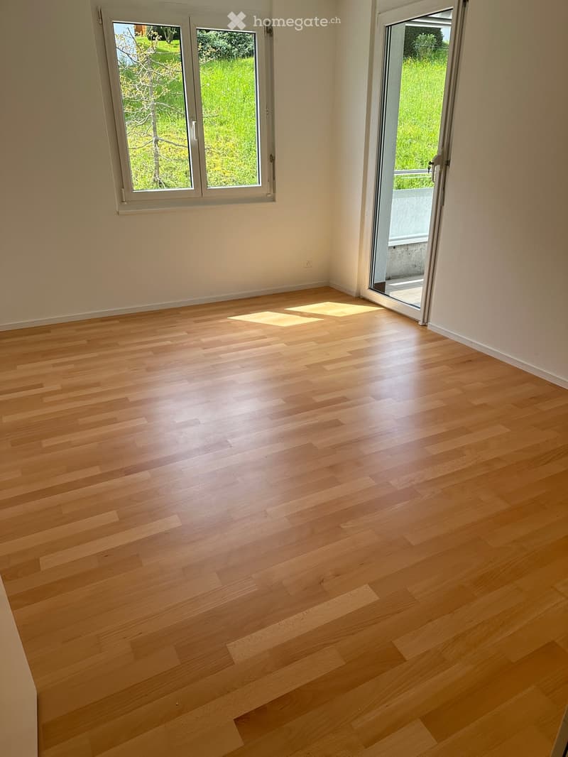 Empty room with light wooden flooring, white walls, and large windows with a view of greenery outside. A sliding glass door leads to a balcony.