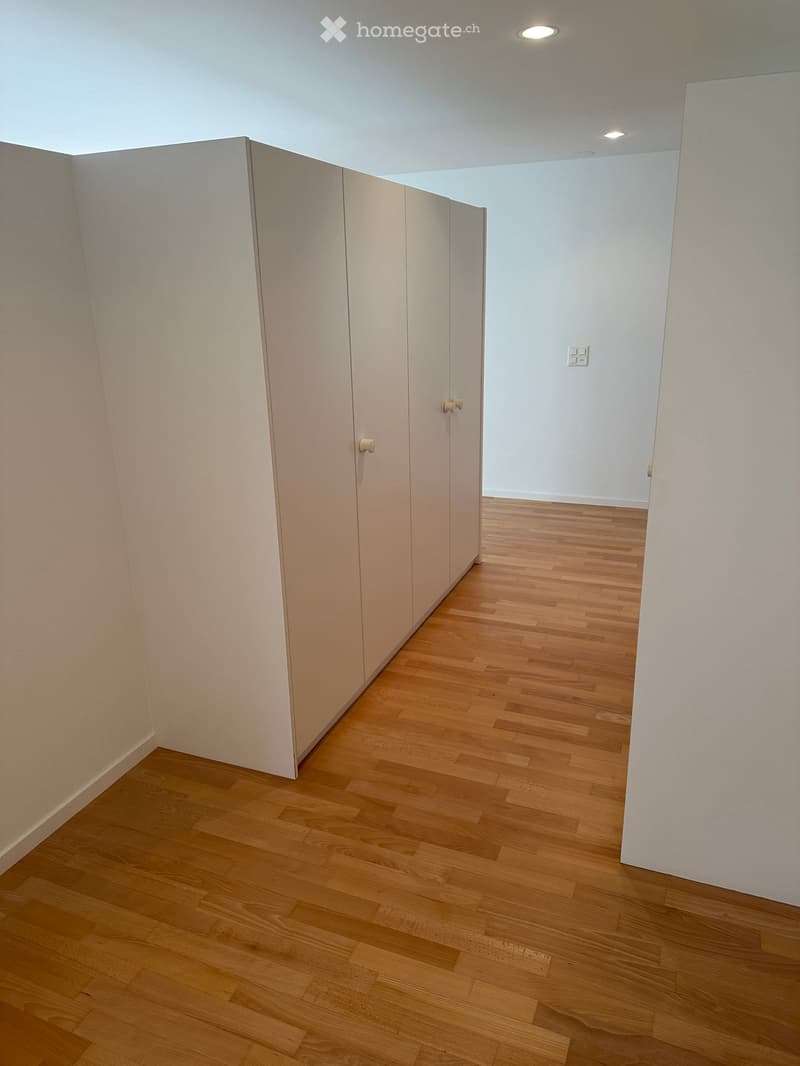Empty hallway with light wooden flooring and white walls, featuring a set of closed white closet doors on one side.