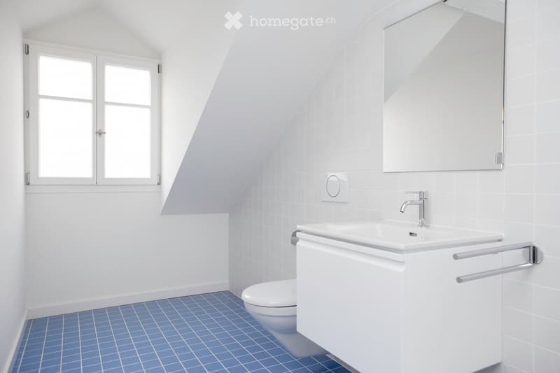 Modern bathroom with white tiles, blue floor tiles, a sink with a modern faucet, a toilet, and a large mirror above the sink. The room features a sloped ceiling and a window with double panes.
