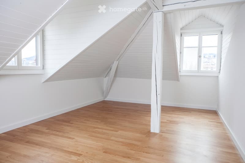 Empty attic room with wooden flooring, white walls, and exposed wooden beams. Two windows provide natural light.