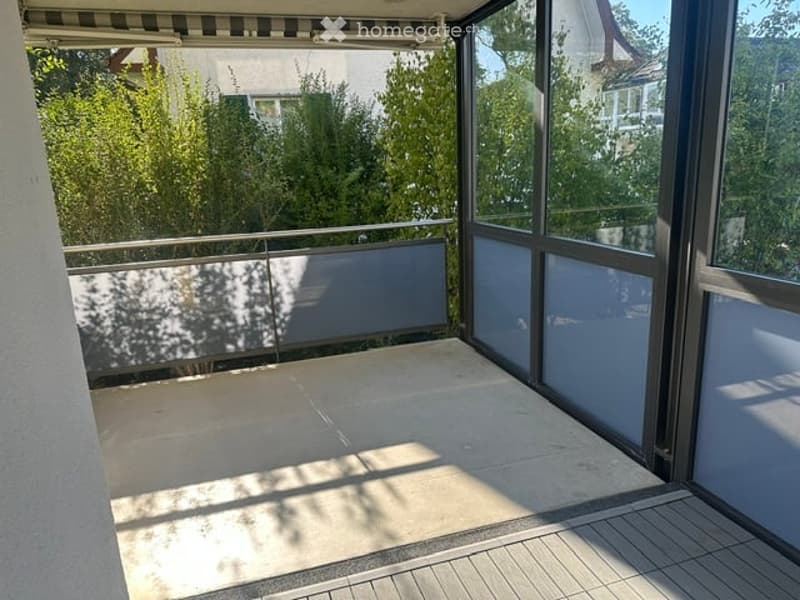 Balcony with large glass panels, metal railing, and tiled floor, overlooking a green area with trees.