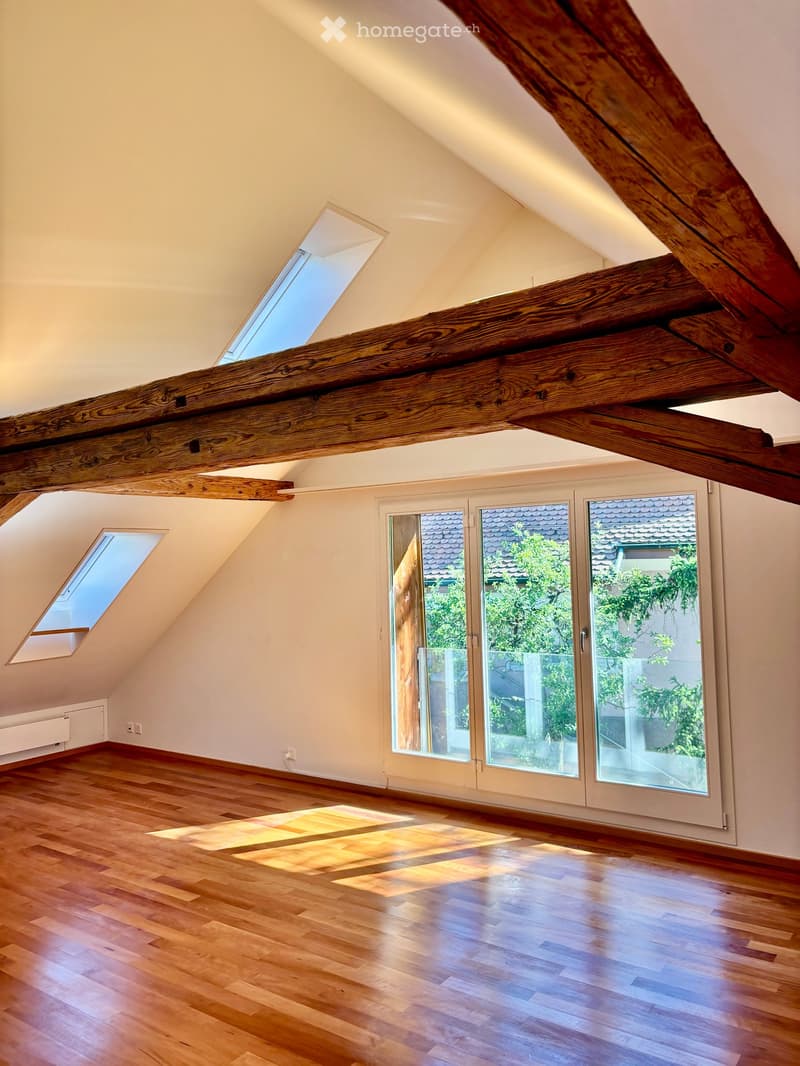 Bright, empty attic room with wooden floors, exposed wooden beams, large windows, and skylights allowing natural light to fill the space.