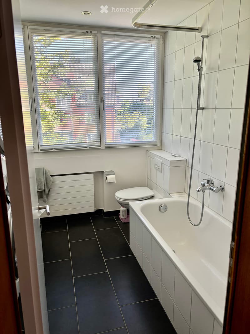 Modern bathroom with white tiles, black tiled floor, bathtub, toilet, radiator, and towel visible.
