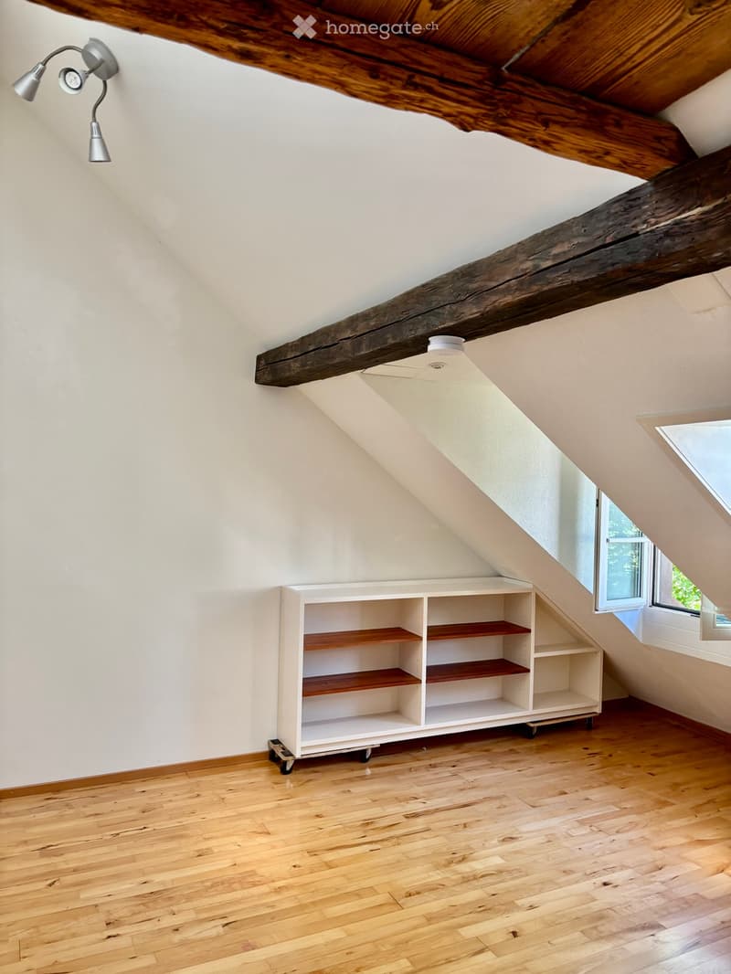 An attic room with wooden beams, exposed wooden ceiling, white walls, hardwood floor, and a built-in white shelving unit with wooden shelves. A small window is visible on the right side.
